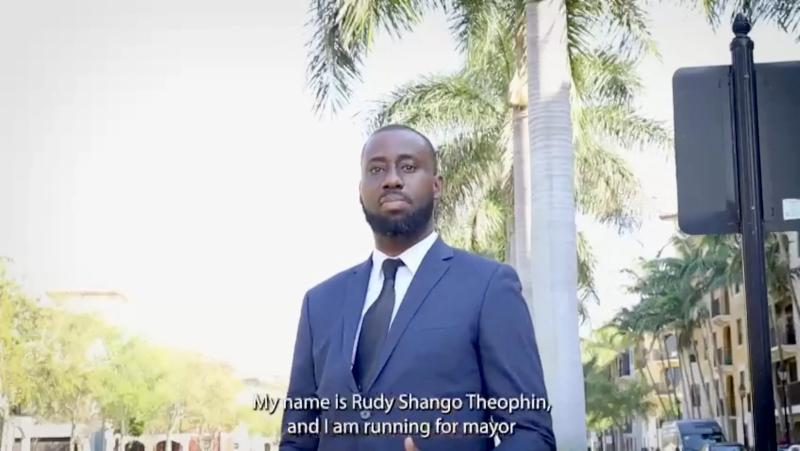 video screenshot of a black man wearing a suit and tie with a caption that reads, "My name is Rudy Shango Theophin, and I am running for mayor."