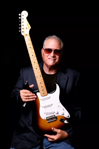 A man with slicked-back hair wearing sunglasses and holding an electric guitar