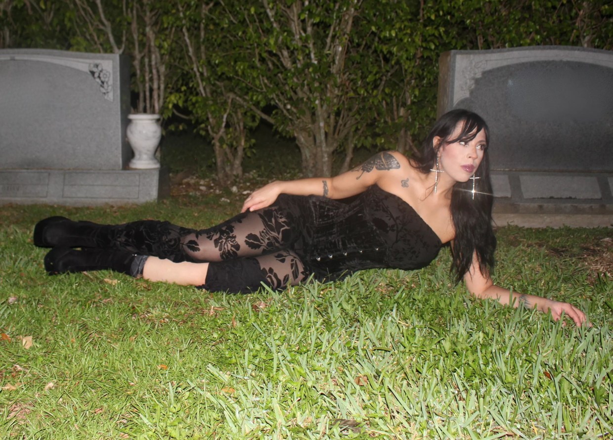 a woman in a black corset and black floral tights lays in the grass at a cemetry