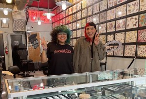 photo of two young women flashing the peace sign inside a tattoo studio with designs on the walls