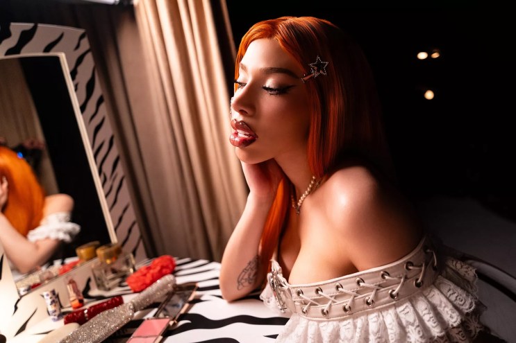 A heavily made-up woman with long red hair, seated at a dressing table