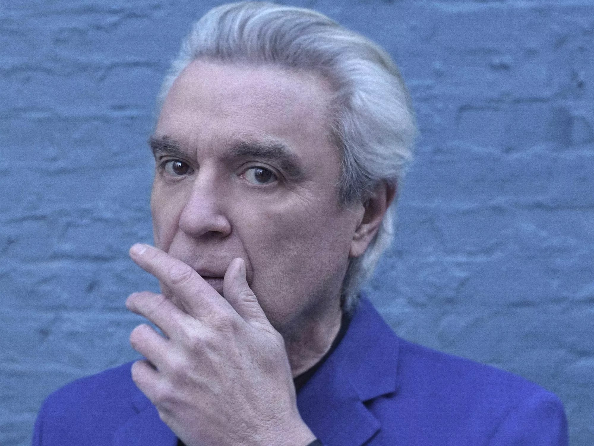 cyan-tinged headshot of David Byrne, with his gray hair swept back and his left hand partially covering his mouth, as if stifling a cough