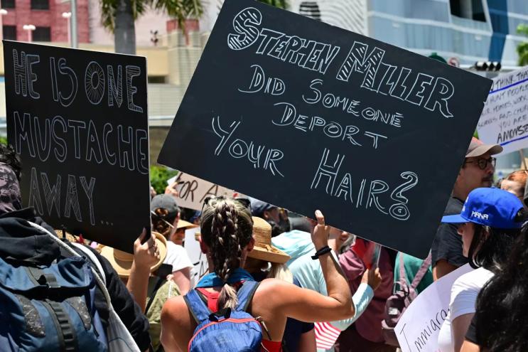 A protester with her back towards the camera holds a black sign that reads, "Stephen Miller did someone deport your hair?"