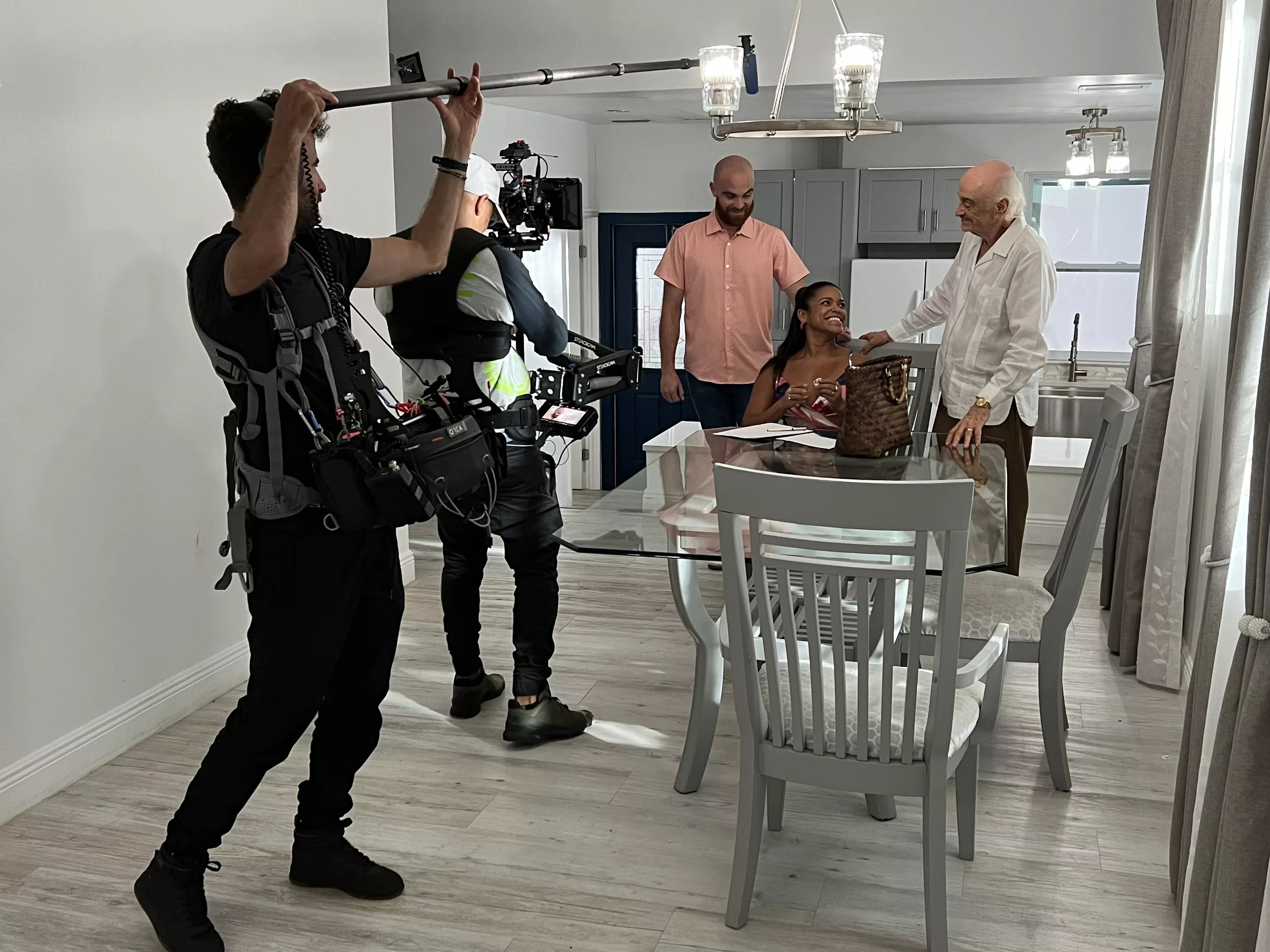 a camera operator and man with a boom mic record an acting scene with two men and a woman at a dining room table