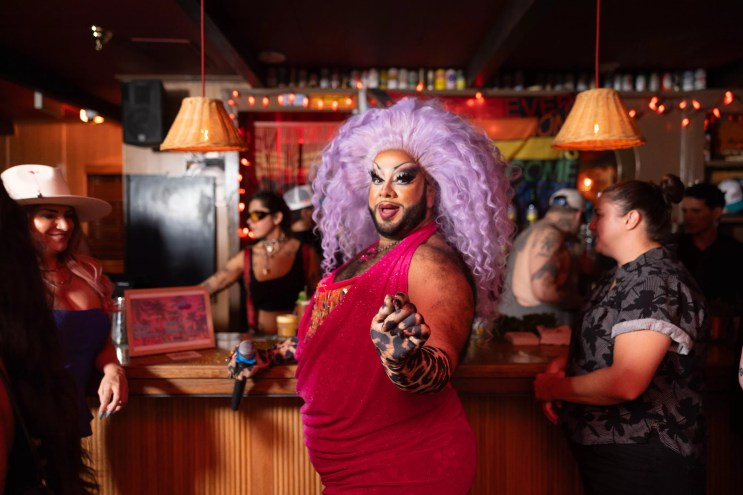 a drag queen in a sparkling pink dress and lavender wig, standing in the middle of a bar